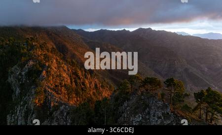 Vista aerea della luce del sole dorata che bacia le cime taglienti e le foreste verdi scure sotto un cielo nuvoloso, Agaete, Isole Canarie, Spagna. Foto Stock