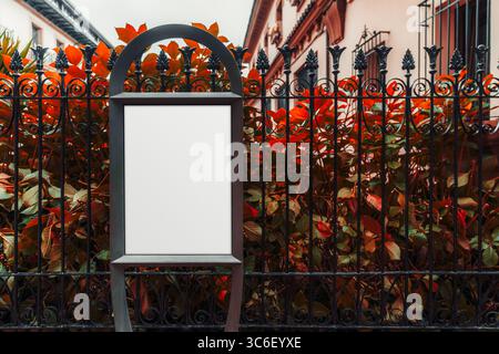 Mockup verticale di tabelloni bianchi con elegante struttura in ferro nero posta davanti a una recinzione in ferro battuto ornata e vivace fogliame rosso, con un tocco classico Foto Stock