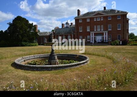 Fontana in pietra in disuso in un prato a Hatchlands Park, con Hatchlands House sullo sfondo, Surrey, Inghilterra, Regno Unito. Scena estiva, 8 luglio 2025. Foto Stock
