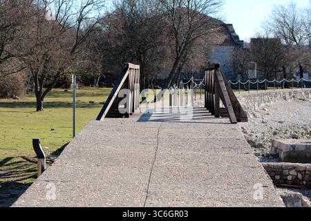 Un solido molo in cemento con corrimano in legno scuro e recinzione in corda conduce da una riva erbosa su lavori in pietra, verso un vecchio edificio, ombreggiato da senza piombo Foto Stock