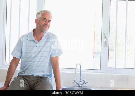 Uomo seduto sul piano di lavoro della cucina piastrellato accanto al rubinetto del lavello in acciaio inox accanto alla finestra, spazio per copiare. Contemporaneo, minimalista, accogliente, informale, naturale Foto Stock