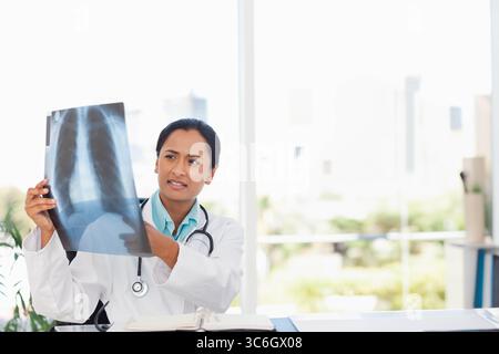Donna indiana medico che esamina la pellicola radiografica del torace in camice bianco da laboratorio alla scrivania della clinica con stetoscopio. Medico, sanitario, diagnostico, professionale, clini Foto Stock