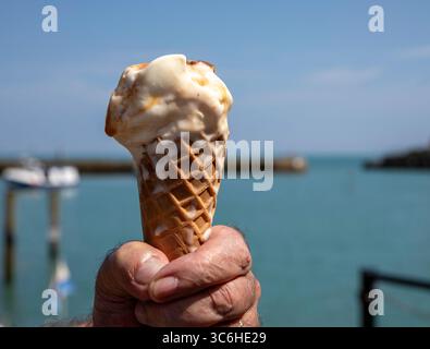 Un uomo regge un cono di waffle che si scioglie in una calda giornata estiva al mare vicino a un porto Foto Stock