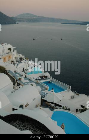Gli iconici edifici bianchi e le piscine di Santorini si affacciano sul Mar Egeo al crepuscolo, con barche che attraversano l'acqua sotto un cielo color pastello. Foto Stock