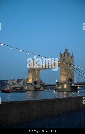 Vista serale del Tower Bridge di Londra illuminato da luci a corda attraverso il cielo, che si riflette nel fiume Tamigi sotto un crepuscolo blu. Foto Stock