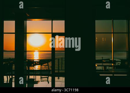 L'enorme tramonto sull'oceano crea una splendida vista attraverso una finestra panoramica. Un vibrante tramonto è incorniciato da finestre vuote di ristorante o caffè Foto Stock