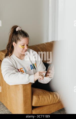 Una donna si siede sul divano con le maschere per gli occhi mentre usa il cellulare Foto Stock
