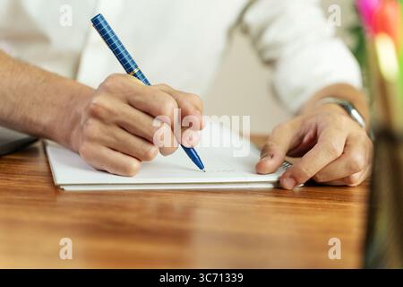 Un dettaglio ravvicinato della mano di un uomo che tiene una penna blu e scrive su un blocco note foderato, prende appunti durante una riunione o firma di un contratto. Foto Stock
