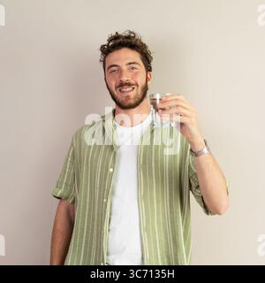 Giovane con una camicia verde che tiene un bicchiere d'acqua, sorride e guarda la macchina fotografica, ambiente interno, stile di vita sano, idratazione, abbigliamento casual, p Foto Stock