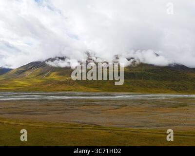 Un'ampia vista aerea di un'aspra montagna con nuvole che rotolano sopra la vetta, che si affaccia su una vasta pianura pianeggiante con un fiume intrecciato Foto Stock