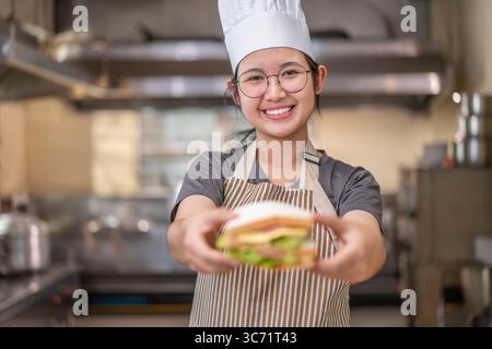 Felice giovane chef asiatica di vent'anni, con occhiali, grembiule e cappello da chef, sorridente e presenta un panino fresco in una cucina commerciale. Ideale Foto Stock