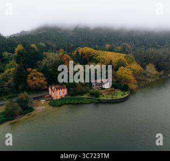 Vista aerea delle case annidate tra il vivace e autunnale fogliame lungo la nebbiosa costa di Lagoa das Furnas, Furnas, Azzorre, Portogallo. Foto Stock