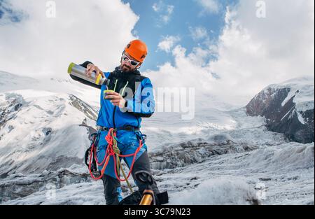 Arrampicatore in attrezzatura da alpinismo sul ghiacciaio durante la salita del picco Lenin sotto le vette della montagna in sottofondo versando una bevanda calda dalla bottiglia termica. AC. Estrema Foto Stock