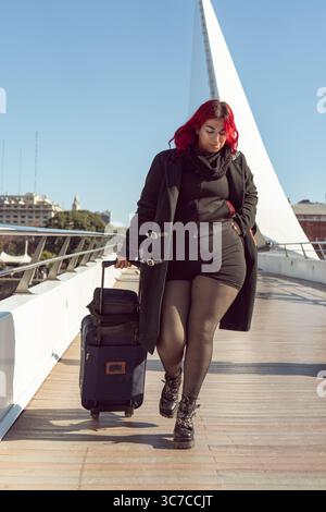 Una donna elegante con i capelli rossi cammina attraverso un ponte a buenos aires, tirando una valigia rotolante dietro di lei Foto Stock