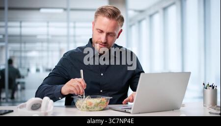 Uomo d'affari sano che mangia insalata alla scrivania mentre lavora su un notebook. Foto Stock