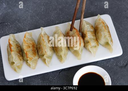 Mano che tiene un paio di bacchette per mangiare gnocchi Mandu o coreani. Accanto al piatto è presente un piccolo recipiente di salsa di soia scura. Foto Stock