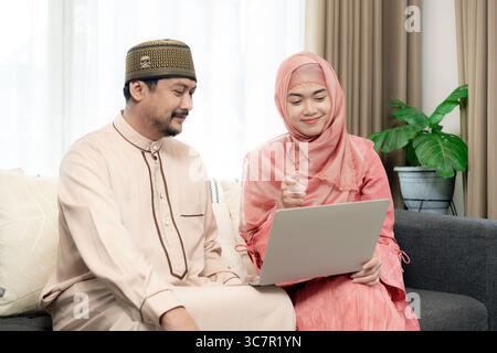 Coppia sorridente che si diverte a fare shopping online a casa, sfogliare i prodotti su un notebook mentre si siede insieme in un salotto accogliente. Foto Stock