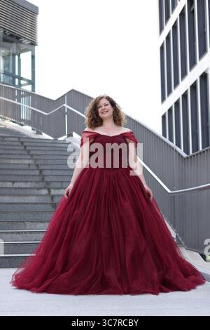 Donna in abito da sera rosso davanti alle scale, sorridente Foto Stock