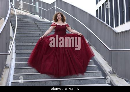 Una donna con un pomposo abito da sera rosso scende le scale, sorridendo Foto Stock