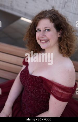 Donna in abito da sera rosso pomposo seduta sulla panchina e sorridente lateralmente Foto Stock