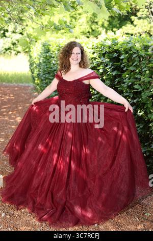 Donna con un pomposo abito da sera rosso davanti a una siepe, vestito disteso Foto Stock