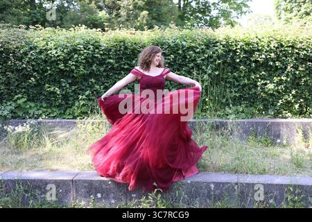 Donna con un pomposo abito da sera rosso si gira di fronte a una siepe Foto Stock
