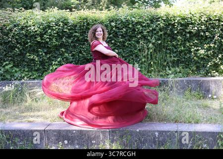 Una donna con un pomposo abito da sera rosso gira più velocemente davanti a una siepe Foto Stock