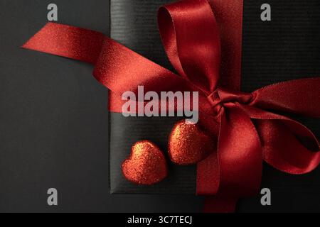 Frammento di una confezione regalo con fiocco rosso e due cuori rossi lucenti su sfondo nero. Regalo per San Valentino. Primo piano. Foto Stock