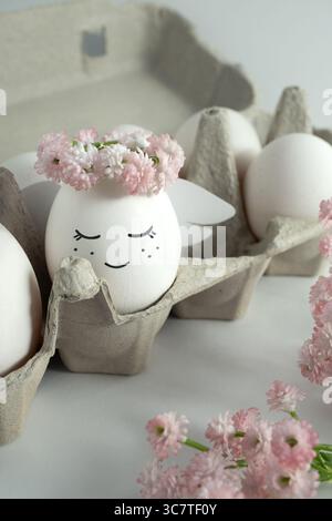 Un angelo fatto di un uovo in una corona di fiori. Scatola delle uova, fiori rosa. Foto Stock
