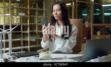 Felice sorridente donna asiatica donna d'affari cinese architetto datore di lavoro che invia messaggi di telefono cellulare scorrimento dei social media addict coreano giapponese Foto Stock