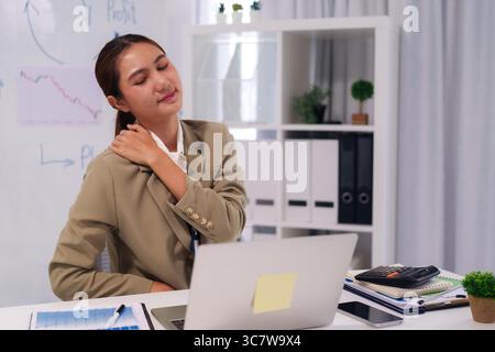 Donna asiatica adulta sperimenta disagio alle spalle in ufficio lavoro lunghe ore. Mostra segni di stress e fatica sul lavoro. Foto Stock