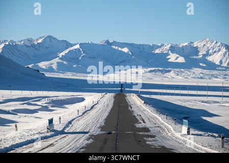 Una strada asfaltata dritta ricoperta di neve e ghiaccio, che conduce verso una vista panoramica delle montagne innevate di Tian Shan sotto un cielo azzurro Foto Stock