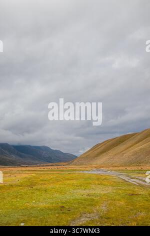 Una strada sterrata si snoda attraverso una vasta valle verde e dorata con una grande collina in pendenza sulla destra e montagne in lontananza sotto un cielo nuvoloso Foto Stock
