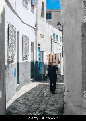L'uomo che trasporta baguette fresche in una scatola di cartone cammina attraverso stretti vicoli nella vecchia medina di Sousse, Tunisia, una scena di routine senza tempo e si Foto Stock