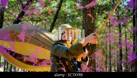Donna che indossa un cappello a secchio e una giacca che stringe la corda nella foresta, con amaca e rivestimento digitale viola Foto Stock