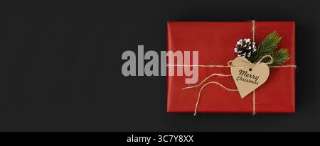 Scatola rossa con etichetta a forma di cuore con il testo Merry christmas, cono e ramoscelli di abete rosso su sfondo scuro. Spazio per il testo yout. Foto Stock
