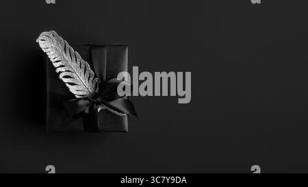Striscione con confezione regalo nera, nastro nero e piuma d'argento su sfondo scuro. Copyspace. Foto Stock