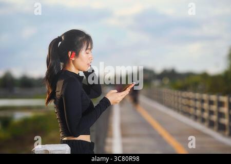 Vista laterale in vita di una giovane donna in piedi su un ponte che indossa auricolari e abbigliamento sportivo che guarda il suo telefono cellulare Foto Stock