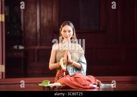Ritratto di una giovane donna che indossa abiti tradizionali thailandesi seduti sul pavimento con offerte religiose in preghiera Foto Stock