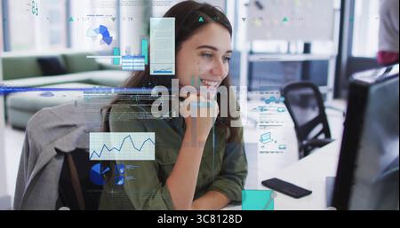 Lavoratore sorridente che esamina i dati sul monitor in ufficio, con rivestimento verde e sovrapposizioni di grafici Foto Stock