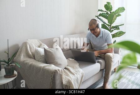 Uomo che lavora da casa e sta videochiamando con un computer portatile e le cuffie mentre beve un caffè Foto Stock