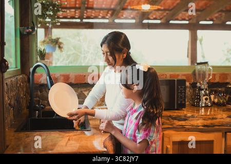La madre sta insegnando a sua figlia come lavare i piatti nella loro cucina rustica, creando un momento commovente di legame familiare e responsabilità condivisa Foto Stock