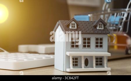 Modello di casa in miniatura sulla scrivania vicino alla calcolatrice e al carrello della spesa, che simboleggia gli investimenti immobiliari, la pianificazione di mutui o l'acquisto di immobili. Foto Stock