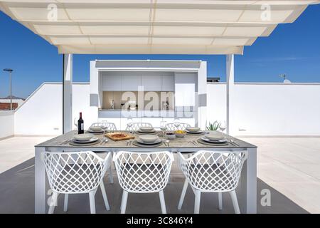 Elegante terrazza sul tetto con moderna cucina all'aperto e tavolo da pranzo completamente allestito sotto un baldacchino. Foto Stock