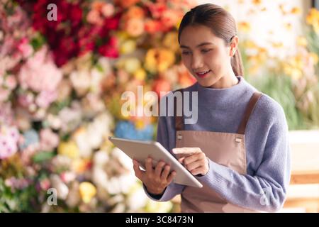 La fioraia donna asiatica adulta gestisce le vendite di scorte tramite tablet digitali migliorando l'esperienza del cliente presso il suo fiorente negozio di fiori Foto Stock