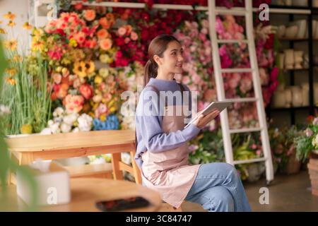 Donna asiatica adulta fiorista lavora felicemente su tablet digitale. L'imprenditore gestisce piccole imprese circondate da vivaci composizioni floreali. Foto Stock