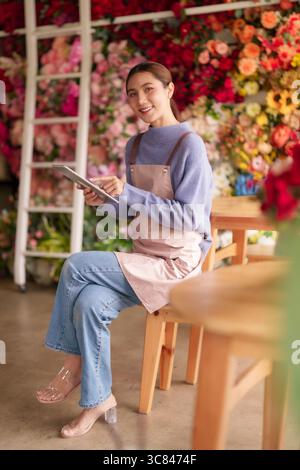 Fioraio donna asiatica gestisce in modo efficiente le attività di fioreria utilizzando la tecnologia dei tablet sorridendo con fiducia tra vivaci esposizioni floreali. Moder Foto Stock