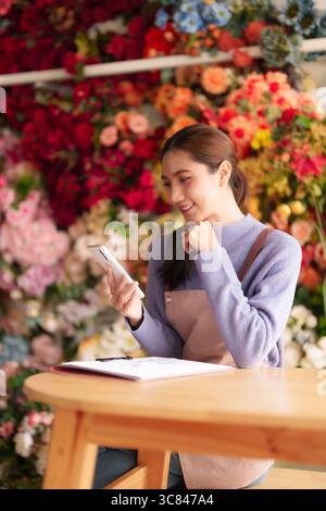 La fioraia donna asiatica gestisce un vivace negozio di fiori con uno smartphone. Gestisce le transazioni aziendali di comunicazione con i clienti e le operazioni quotidiane. Foto Stock