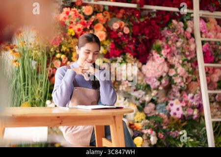 Un'affascinante giovane donna asiatica fioraia lavora diligentemente nel suo vivace negozio di fiori. Sorride durante la pianificazione delle operazioni quotidiane e la gestione degli ordini Foto Stock