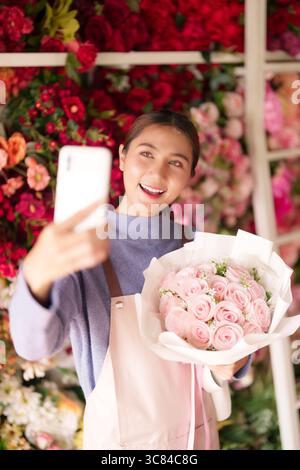 Il gioioso fiorista asiatico cattura splendidi momenti di bouquet di rose rosa utilizzando lo smartphone nel suo vivace negozio di fiori che mostra arte floreale. Foto Stock